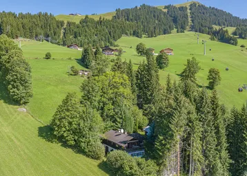 Luftaufnahme eines Hauses in einer grünen Berglandschaft mit vielen Bäumen und Skipisten.