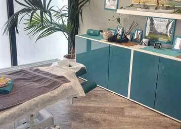Behandlungsraum mit Massageliege, modernen teal-farbenen Schränken und Holzoptikboden, hell durch großes Fenster.