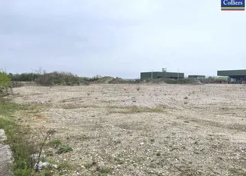 Großes, unbebautes Grundstück mit Kiesboden und spärlicher Vegetation, im Hintergrund sind Industriegebäude sichtbar.