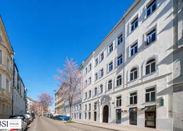 Helle Fassade eines gepflegten Mehrfamilienhauses in einer städtischen Straße mit blühenden Bäumen im Frühling.