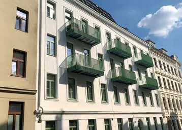 Weißes Mehrfamilienhaus mit grünen Balkonen und Fenstern unter blauem Himmel.