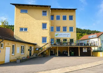 Gelbes Mehrfamilienhaus mit mehreren Etagen und Parkplätzen im Vordergrund, umgeben von grünen Hügeln.