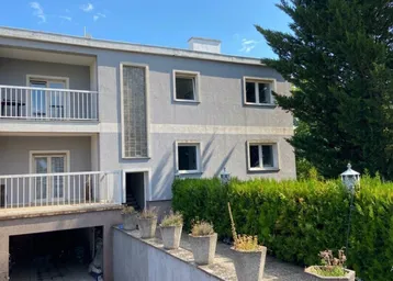 Graues Mehrfamilienhaus mit zwei Balkonen und einer Garageneinfahrt, umgeben von grünen Hecken.