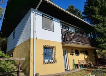Zweistöckiges Haus mit gelb-weißer Fassade, Holzbalkon und umgebender Natur.