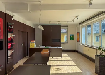 Geräumiges Büro mit großen Fenstern, Holzboden und modernen Möbeln in dunklen Farbtönen, gut beleuchtet.