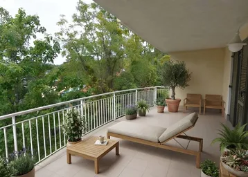 Geräumiger Balkon mit bequemer Sonnenliege, kleinem Tisch und üppiger Bepflanzung, umgeben von grünen Bäumen.