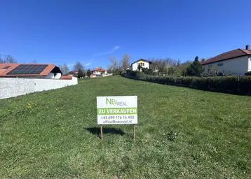 Grünes Grundstück mit 'Zu verkaufen'-Schild und umliegenden Häusern unter blauem Himmel.