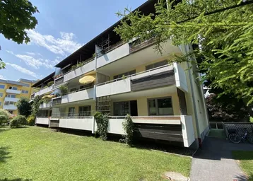 Mehrfamilienhaus mit mehreren Balkonen und gepflegter Grünfläche im Vordergrund unter blauem Himmel.