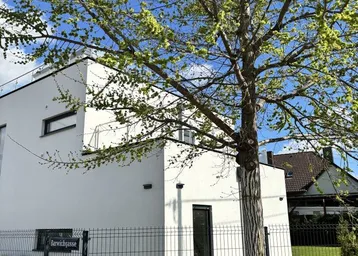 Modernes weißes Gebäude mit klaren Linien und einem Baum im Vordergrund unter blauem Himmel.