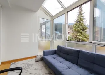 Heller Wintergarten mit blauer Couch, Glasdach und Bergblick durch große Fenster.