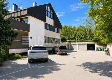 Mehrfamilienhaus mit Parkplätzen und Garagen in grüner Umgebung unter blauem Himmel.