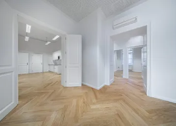 Geräumiger, heller Raum mit Fischgrätparkett, weißen Wänden und einer kleinen Küchenzeile im Hintergrund.