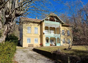 Historische Villa mit gelber Fassade und grünem Balkon, umgeben von Bäumen unter blauem Himmel.