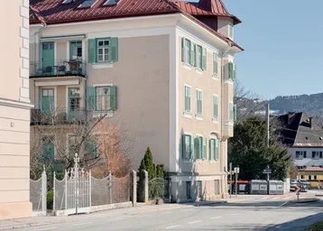 Historisches Gebäude mit beiger Fassade, roten Dachziegeln und grünen Fensterläden an einer Straße.