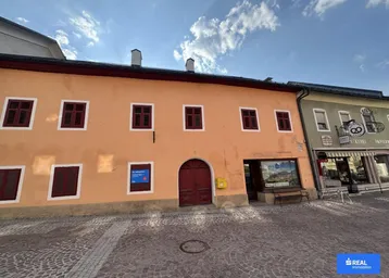 Historisches Gebäude mit orangefarbener Fassade, roten Fensterläden und Geschäften im Erdgeschoss an einer Kopfsteinpflasterstraße.