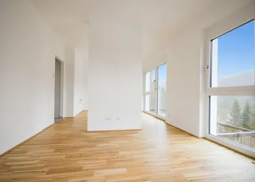 Helles, leeres Zimmer mit Holzboden und großen Fenstern, die einen Blick ins Grüne bieten.