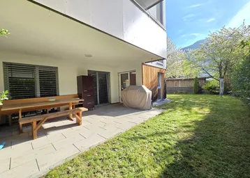 Geräumige Terrasse mit Holztisch und Gartenbereich, umgeben von grüner Hecke und Bergblick.