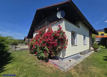 Hausfassade mit blühenden Rosensträuchern und gepflegtem Rasen unter strahlend blauem Himmel.