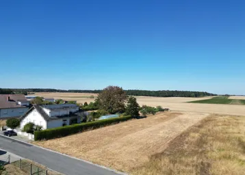 Ländliche Umgebung mit weiten Feldern, einigen Wohnhäusern und einer Hecke unter klarem Himmel.
