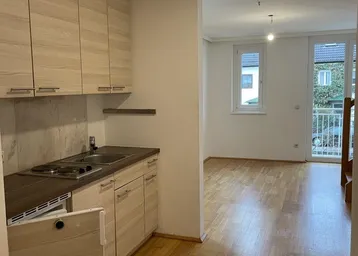 Helle offene Wohnfläche mit integrierter Küchenzeile, Parkettboden und Zugang zu einem Balkon.