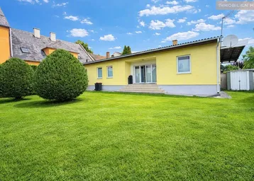 Großer, gepflegter Garten mit Rasenfläche und Sträuchern hinter einem gelben Gebäude unter blauem Himmel.