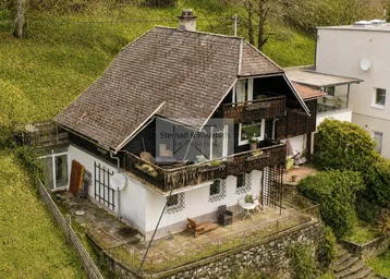 Traditionelles Haus mit Holzbalkonen und einer möblierten Terrasse in Hanglage, umgeben von viel Grün.