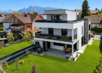 Modernes Wohnhaus mit Flachdach, großem Garten und Bergblick unter blauem Himmel.