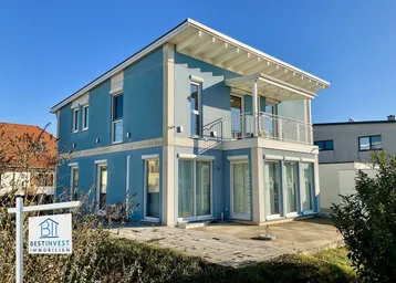 Modernes Einfamilienhaus mit blauer Fassade, Balkon und großen Fenstern unter blauem Himmel.