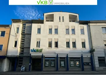 Mehrstöckiges Gebäude mit weißer Fassade und Geschäftsräumen im Erdgeschoss an einer Straße.