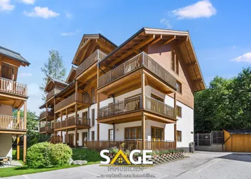 Modernes Mehrfamilienhaus im alpinen Stil mit Holzfassade, weißen Wänden und zahlreichen Balkonen, umgeben von Grünflächen.