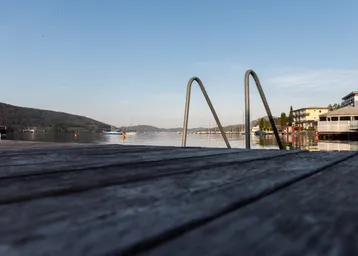 Holzsteg mit Badeleiter am Seeufer, umgeben von Bergen und Gebäuden, mit Blick auf Segelboote.