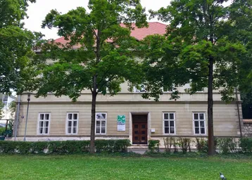 Historisches Gebäude mit klassischer Fassade, umgeben von altem Baumbestand und einer gepflegten Rasenfläche.