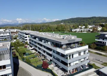 Luftaufnahme moderner Wohngebäude mit Flachdächern, umgeben von Grünflächen und Bergen im Hintergrund.