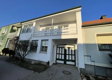Weißes Gebäude mit grünem Garagentor und einem Balkon im Obergeschoss, umgeben von einer kleinen Gartenfläche.