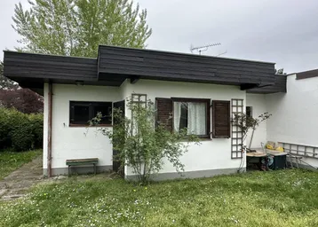 Kleines Haus mit weißer Fassade, dunklem Flachdach und grünem Vorgarten.