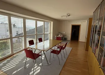 Heller Büroraum mit großem Holztisch, roten Stühlen und breiten Fenstern, die viel Tageslicht hereinlassen.