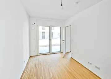 Helles, leeres Zimmer mit Holzboden und großen Fenstern zum Außenbereich.