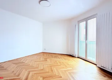 Helles Zimmer mit Fischgrätparkettboden, weißen Wänden und Zugang zu einem französischen Balkon.