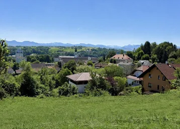 Panoramablick über ein grünes Dorf mit Häusern, Bäumen und einer Kirche im Hintergrund, umgeben von Bergen.