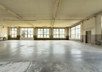 Großer, leerer Raum mit Betonboden und vielen Fenstern, ideal für Lager oder Produktion.