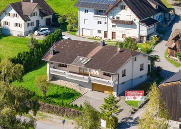Luftaufnahme eines Einfamilienhauses mit braunem Dach und umliegenden Grünflächen in einer Wohnsiedlung.