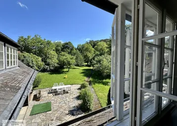 Blick aus dem Fenster auf eine weitläufige Gartenanlage mit Steinterrasse und Sitzgelegenheiten.