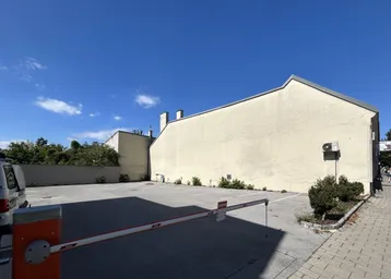 Außenansicht eines Gebäudes mit angrenzendem Parkplatz und Zufahrtsschranke unter blauem Himmel.