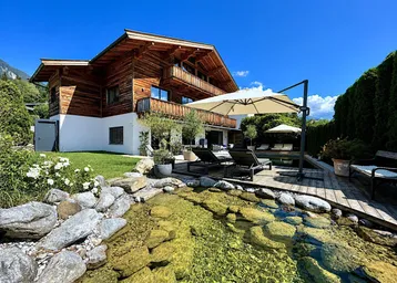Charmantes Holzchalet mit gepflegtem Garten, Teich und Sonnenterrasse vor alpiner Bergkulisse.