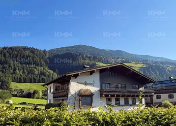 Traditionelles Haus im alpinen Stil mit Holzbalkonen und Bergblick, umgeben von grünen Hügeln und Wäldern.