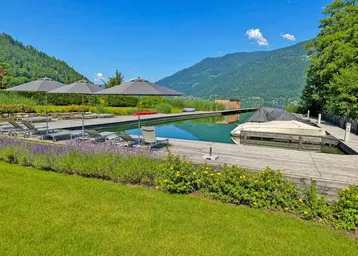 Großer Naturpool mit Holzdeck, Liegestühlen und Blick auf den See und die Berge.