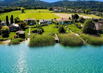 Luftaufnahme von Häusern am Ufer eines klaren Sees, umgeben von grünen Wiesen und bewaldeten Hügeln.