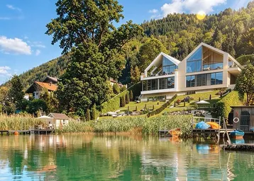 Moderne Villa am Seeufer mit weitläufigem Garten und Bergblick unter strahlend blauem Himmel.