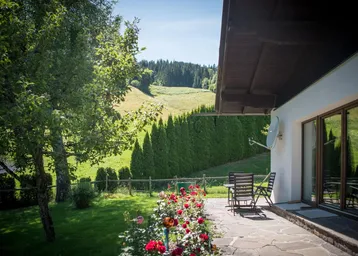 Sonnige Terrasse mit Steinplatten, Gartenmöbeln und Blick auf grüne Hügel und Wald.