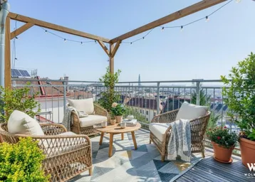Sonnige Dachterrasse mit gemütlicher Rattan-Sitzgruppe, Holzpergola, Lichterkette und weitem Stadtblick.